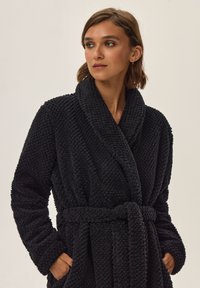 Czarna szlafrok z polaru o teksturowanym wzorze, zapinany na pasek, z szerokim kołnierzem i wiązaniem w talii. Posiada dwie boczne kieszenie dla wygody.