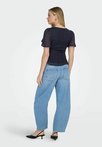 Femme de profil portant un haut en dentelle bleu marine à manches courtes, un jean ample bleu clair et des chaussures à talons bas noires à bride arrière.