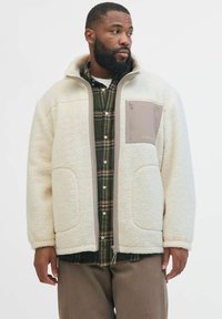 Giacca in pile di colore bianco sporco con colletto alto, zip frontale e tasca sul petto beige, indossata sopra una camicia in flanella a quadri. Tessuto morbido e strutturato.