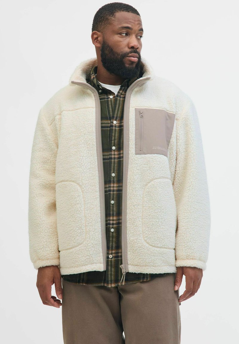 Giacca in pile di colore bianco sporco con colletto alto, zip frontale e tasca sul petto beige, indossata sopra una camicia in flanella a quadri. Tessuto morbido e strutturato.
