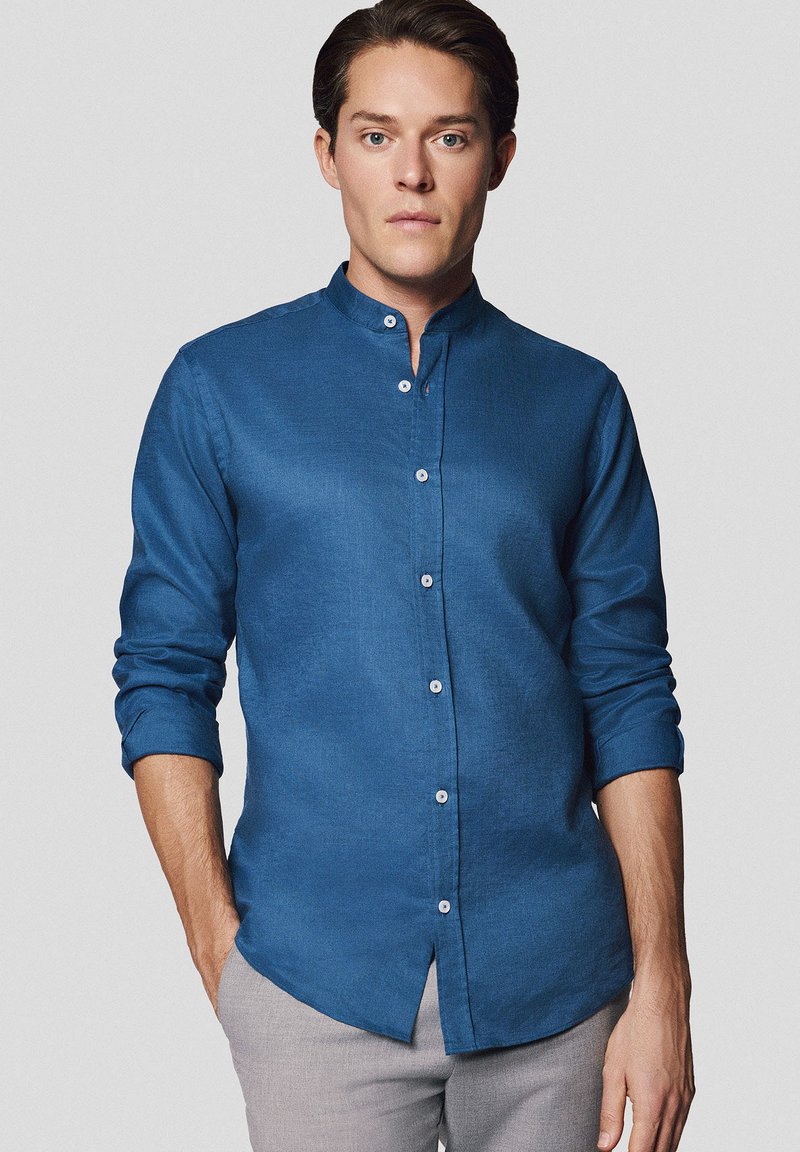 Niebieska, długa koszula z rękawami, z kołnierzykiem mandaryńskim, wykonana z fakturowanej tkaniny, posiadająca guziki z przodu i luźny krój.