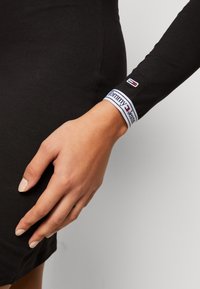 Vestido negro de manga larga con diseño ajustado, que presenta un puño a rayas blancas y marinas con la marca "Tommy Jeans". Textura de tela suave.