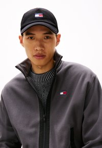 Grå fleecejakke med sorte detaljer, lynlås-lukning og lille Tommy Jeans-logo. Sort kasket med et flerfarvet logo-patch foran.