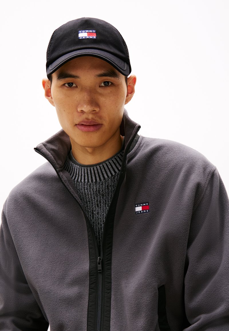 Veste en polaire grise avec des accents noirs, fermeture zippée et petit logo Tommy Jeans. Casquette noire avec un patch logo multicolore à l'avant.