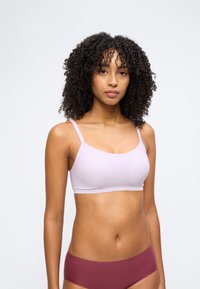 Γυναίκα με σγουρά μαλλιά που φοράει ανοιχτό μοβ σουτιέν και σκουρόχρωμο μπορντό εσώρουχο, στέκεται μπροστά από απλό ανοιχτό γκρι φόντο.