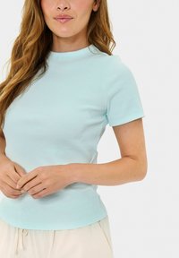 Světle modrý žebrovaný top s krátkým rukávem a kulatým výstřihem, doplněný o neutrální béžové kalhoty. Materiál vypadá měkce a texturovaně.