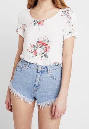 Kobieta w białej bluzce w kwiaty z krótkim rękawem, wpuszczonej w jasnoniebieskie, podarte szorty jeansowe z postrzępionymi krawędziami.