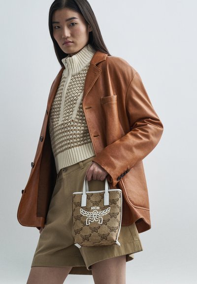 Kobieta w brązowej skórzanej marynarce, beżowym swetrze dzianinowym i dopasowanych szortach, trzymająca małą torebkę MCM w wzór z białymi uchwytami.