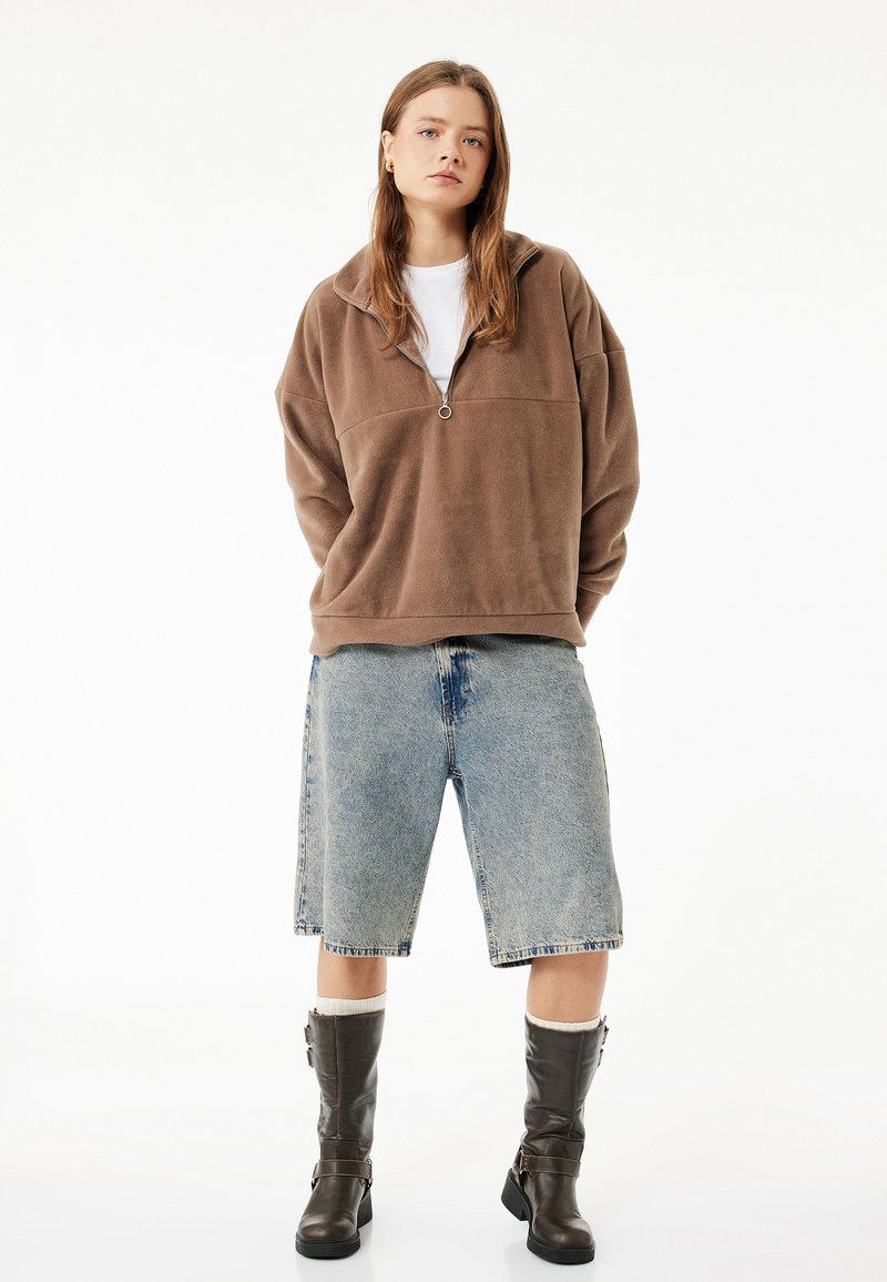Brun fleece pullover med halv lynlås, lyseblå denim bermudashorts og knæhøje brune støvler med spænder. Hvid skjorte under.