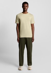 Gaiši bēša īsām piedurknēm T-krekls ar apaļu apkakli, kombinēts ar olīvu zaļām biksēm. Uz krūtīm redzama neliela logo detaļa.