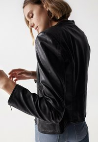 Schwarze Lederjacke mit tailliertem Schnitt, sichtbaren Steppnähten und Reißverschlusstaschen. Glatte Textur und angepasste Ärmel.