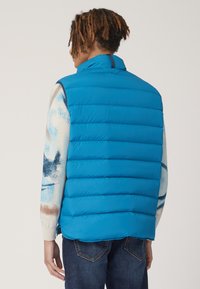 Gilet blu imbottito con design a righe verticali, colletto alto e una texture liscia e lucida. Indossato sopra un maglione a fantasia e jeans scuri.