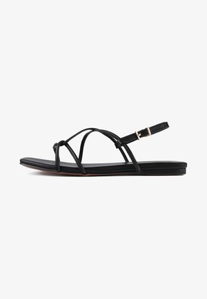 Sandales plates noires avec un design de lanière nouée, boucle de cheville ajustable, texture en cuir lisse et silhouette minimaliste.