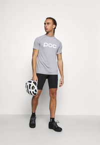 Grå atletisk t-shirt med "poc" logo, parret med sorte shorts og sko. Bæreren holder en hvid hjelm med sorte accenter.