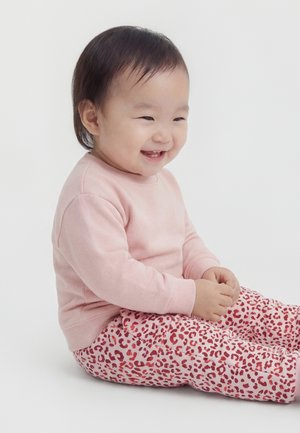 Bambino seduto e sorridente, indossa un maglione rosa chiaro e pantaloni rosa con motivo leopardato rosso su sfondo bianco.