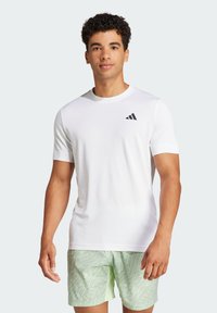Camiseta deportiva blanca con mangas cortas y escote redondeado. Detalle del logo negro en el lado izquierdo del pecho. Tejido mate y suave.