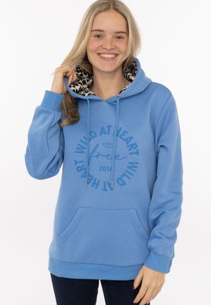 Young woman with long blonde hair smiling, wearing a blue hoodie with "Wild at heart free" text and animal print lining in the hood.