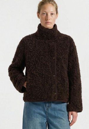 Femme portant une veste en polaire brun foncé texturée avec un col montant et un jean bleu, debout devant un fond clair uni.