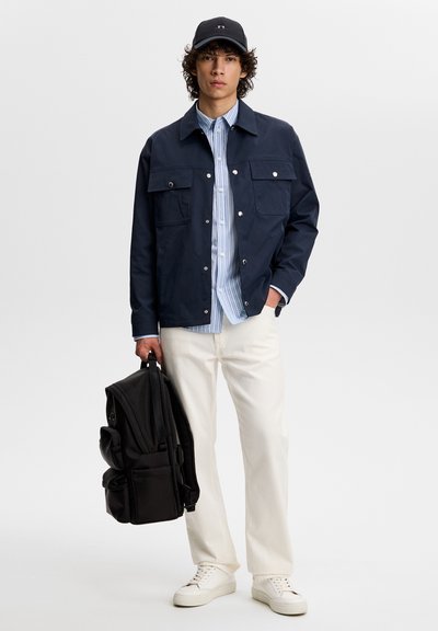 Giovane uomo con giacca blu navy, camicia a righe blu, pantaloni bianchi, sneakers bianche, berretto nero, che tiene uno zaino nero di lato.