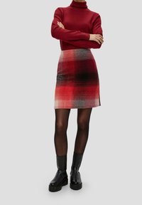 Personne portant un pull col roulé rouge, une jupe à carreaux rouge et noir, des collants noirs transparents et des bottines à plateforme noires, se tenait les bras croisés.