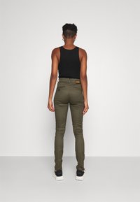 Svart linne, olivgröna figursydda byxor med subtil textur, tre bakfickor och en etikett sydd i midjan. Avslappnade skor synliga.