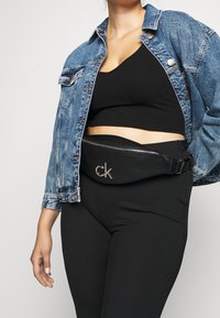 Černá kožená taška na pás se stříbrnými detaily, se zipem a embosovaným logem, nošena přes černý žebrovaný crop top a džínovou bundu.
