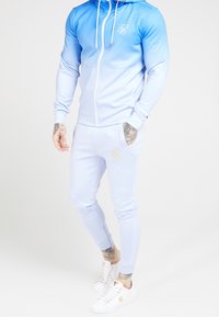Felpa azzurra chiara e pantaloni coordinati in un tessuto liscio con effetto ombre, con un logo sul petto e sul lato dei pantaloni.