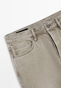 Licht beige spijkerbroek met voorzak, riemlussen en metalen knoopsluiting op een witte achtergrond.