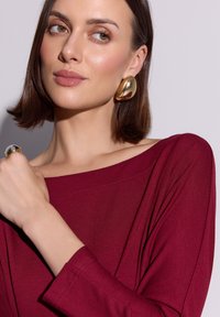 Bordowa bluzka z szerokim dekoltem i długimi rękawami, zestawiona ze złotymi kolczykami wiszącymi i metalicznym pierścionkiem, gładka faktura tkaniny.