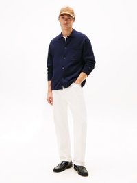 Camicia blu navy con colletto, tasca sul petto, jeans bianchi e scarpe nere. Cappello color corda con un dettaglio del logo.