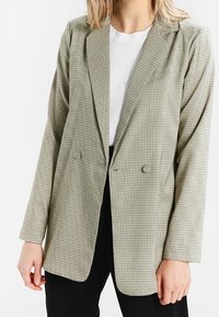 Zielona i biała marynarka w kratę z klapami z wycięciem, zapinana na podwójne guziki, z długimi rękawami. Materiał wydaje się mieć fakturę.