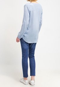 Camicia a maniche lunghe azzurro chiaro con pois, abbinata a jeans scuri e sneaker bianche. La camicia presenta un orlo arrotondato e polsini con bottoni.