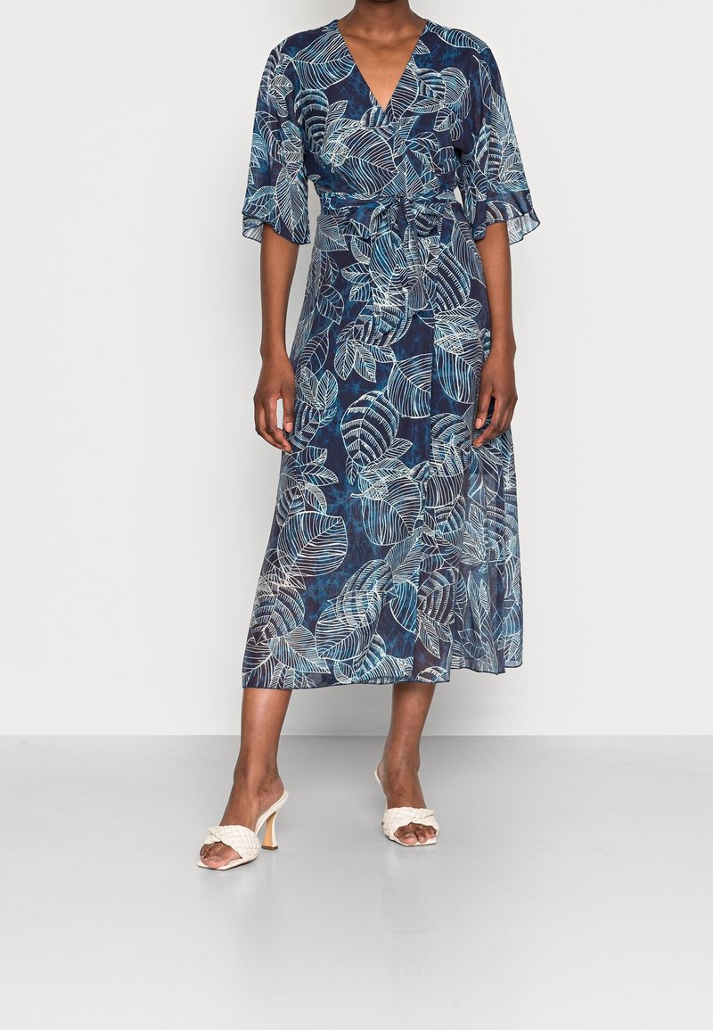 Femme portant une robe midi portefeuille bleu marine avec des motifs de feuilles blanches et des manches évasées, associée à des sandales blanches tressées à talons.