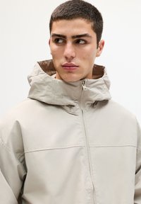 Veste imperméable beige clair avec col haut, fermeture éclair et poignets élastiques. Texture lisse et design minimaliste sans logos visibles.