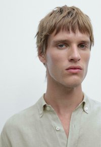 Lysbeige linned skjorte med knap-nyt krave, der har en afslappet pasform. Modellen har kort, ustyrligt blond hår og en neutral baggrund.