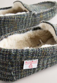 Chaussons doux, doublés en polaire, avec extérieur en tissu Harris Tweed gris-vert et une étiquette de marque visible sur le côté.