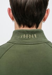 Pull-over vert olive à col montant, avec le logo "JORDAN" brodé en or dans le dos. Tissu doux et texturé avec une coupe ajustée.
