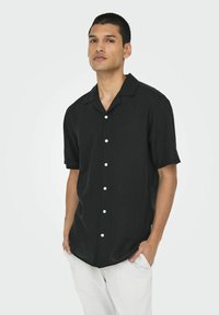 Camisa de botones de manga corta negra con cuello, con botones blancos y un corte relajado, hecha de una tela texturizada.