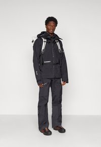 Casaco e calças impermeáveis pretos com fechos, punhos ajustáveis e acentos de logotipo; calçado robusto com solas texturizadas e padrões coloridos.