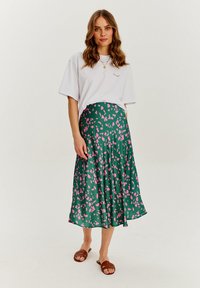 Zielona spódnica w wzory z różowymi kształtami liści, połączona z białym, dużym t-shirtem i brązowymi sandałami. Gładki materiał, średniej długości fason.