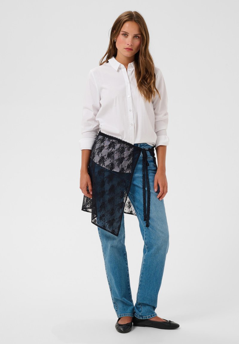 Femme debout portant une chemise blanche boutonnée, un jean bleu, une jupe portefeuille en dentelle noire nouée à la taille, et des ballerines noires sur fond blanc.
