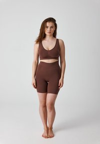 Woman standing barefoot wearing brown seamless sports bra and high-waisted biker shorts against plain light background.