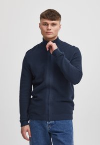 Granatowa prążkowana kurtka na zamek z wysokim kołnierzem, charakteryzująca się gładkim, teksturowanym wykończeniem oraz kontrastującym pionowym paskiem.