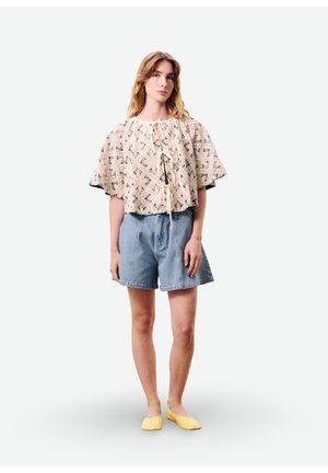 Femme portant un haut ample beige à motifs avec des liens, un short taille haute bleu et des ballerines jaunes, debout devant un fond uni.