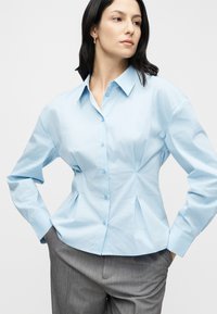 Chemise bleu clair à boutons avec un design structuré, des côtés plissés, un col pointu et des manches longues. Portée avec un pantalon gris.