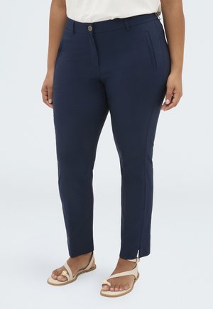 Personne portant un pantalon bleu marine slim ajusté aux chevilles avec une fente latérale et des sandales beige à bout ouvert, debout devant un fond clair uni.