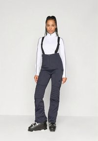 Marineblå skioverall med justerbare svarte seler, ribbestruktur på lårene, stylet med en hvit langermet topp med høy hals.