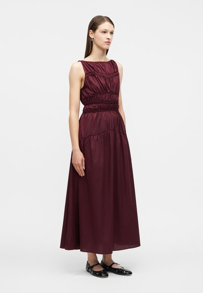 Γυναίκα που στέκεται φορώντας αμάνικο, ρυτιδωτό μακρύ φόρεμα σε μπορντό απόχρωση και μαύρα επίπεδα παπούτσια με μεταλλικές τρύπες, με φόντο μια απλή επιφάνεια.