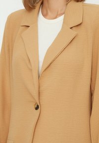 Blazer marrón claro hecho de tela texturizada, con solapa muesca, cierre de un solo botón, y usado sobre una blusa blanca.