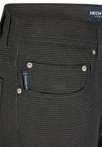 Pantalons noirs à motifs avec un tissu texturé, présentant une petite poche avant et des accents métalliques sur les poches arrière. Étiquette visible.